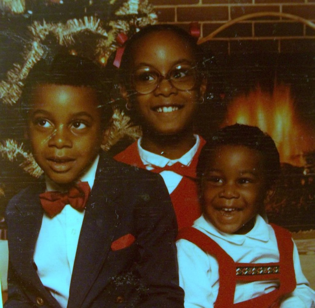 Drei Kinder lachen vor einem Weihnachtsbaum mit einem Kamin aus Holzscheiten auf der rechten Seite.