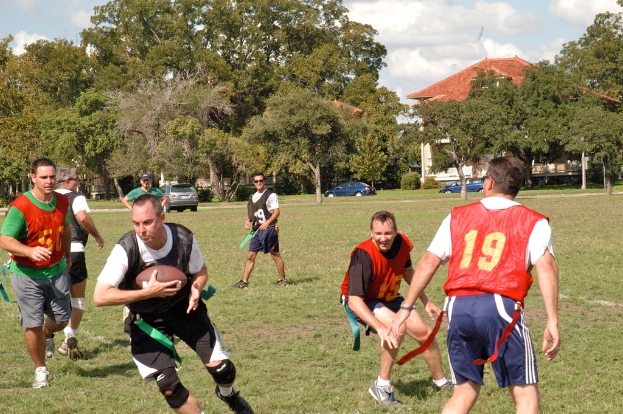 Leute, die Amerikanischen Fußball auf einem grünen Feld mit einem Haus, Autos und Bäumen im Hintergrund spielen.