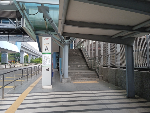 Ein Bahnsteig mit Treppe, eine Straße mit Geländern auf der linken Seite und ein Hintergrund mit Bäumen, Pfählen, Gebäuden und einer Brücke, mit Rohren und einer Tafel am oberen Ende des Bahnsteigs.