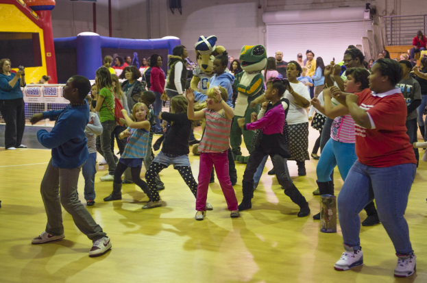 Kinder tanzen in einer Turnhalle mit einer Maskottchenfigur, umgeben von Zuschauern mit Kameras und aufblasbaren Gegenständen im Hintergrund.