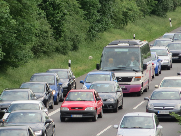 Ein Stau auf einer Autobahn mit vielen Autos und einem Lieferwagen, Menschen sind in den Fahrzeugen sichtbar, Bäume und Gras im Hintergrund.