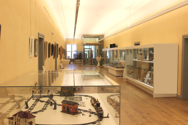 Ein unordentlicher Raum mit verschiedenen Spielzeugen, darunter ein Spielzeugholzset auf einem Glastisch, gerahmte Fotos an den Wänden, Schränke mit Gegenständen darin, eine Tür, ein weiterer Tisch mit Gegenständen darauf und Deckenleuchten.