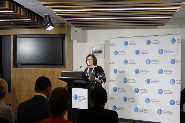 Eine Frau steht auf einer Bühne und spricht in ein Mikrofon, vor ihr sitzen Zuschauer, an der Wand hinter ihr eine Fahne, ein Fernseher an der hölzernen Wand und Lampen an der Decke.