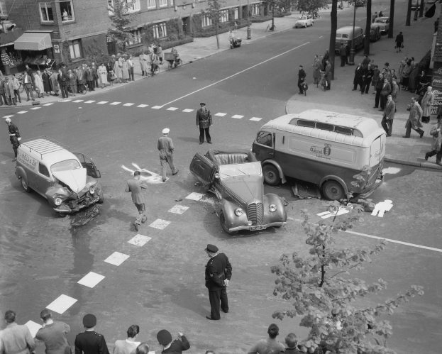 Schwarz-weiß-Szene eines Autounfalls an einer Straßenkreuzung mit mehreren Fahrzeugen, darunter ein Van, und einer Gruppe Schaulustiger; Gebäude, Bäume, Pfähle und Texttafeln sind im Hintergrund zu sehen.