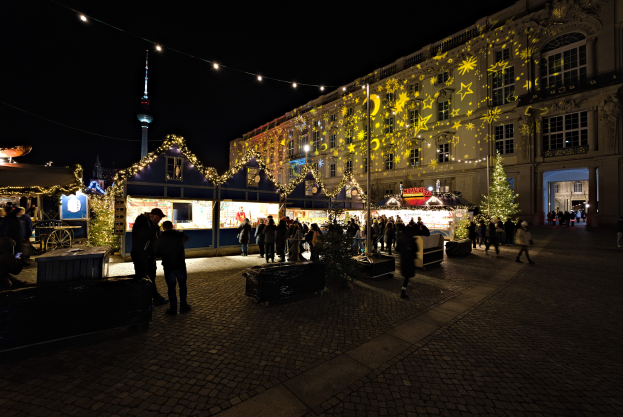 Ein lebendiger Weihnachtsmarkt in Berlin, Deutschland, mit Menschen um beleuchtete Stände, festliche Décoration und Gebäude mit beleuchteten Fenstern unter einem dunklen Himmel.
