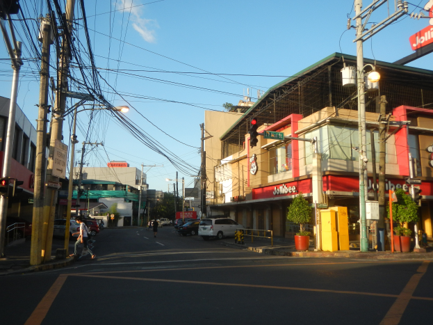 Eine Stadtstraße mit Autos, Fußgängern, Strommasten, Verkehrszeichen, Gebäuden mit Schildern, Topfpflanzen und einer bewölkten Himmel.