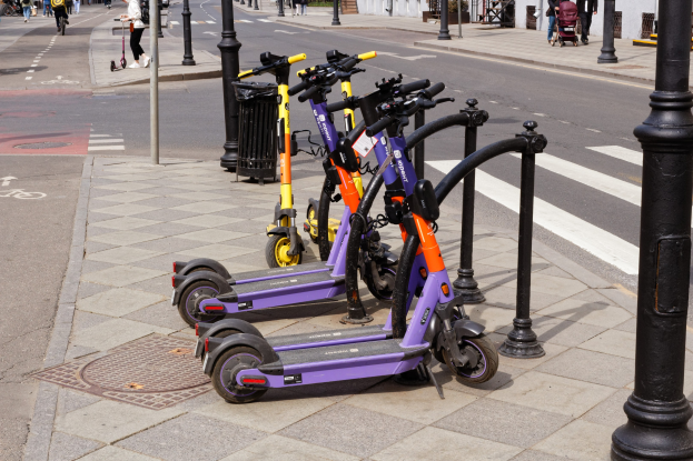 Eine Reihe von Elektro-Scootern, die auf der Seite einer Straße geparkt sind, mit Menschen, die auf dem Gehweg gehen und Fahrräder fahren, einem Müllcontainer im Vordergrund und Gebäuden, Bäumen und Pflanzen im Hintergrund.