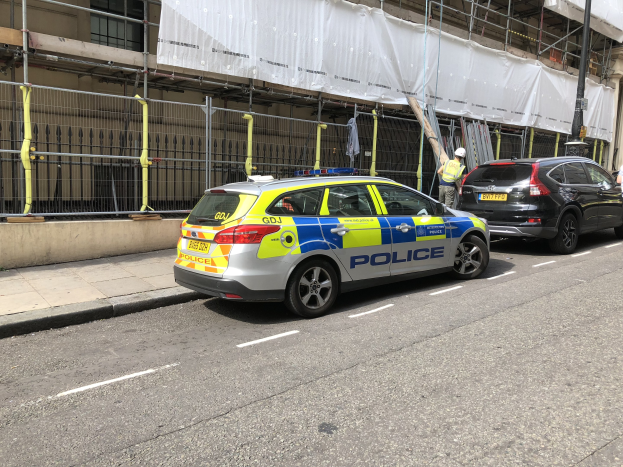Polizeiauto auf einer Straße neben einem Gebäude geparkt, mit zwei Helikopterpiloten und einem weißen Tuch über dem Gebäude, wahrscheinlich nach einer Messerstecherei in London.