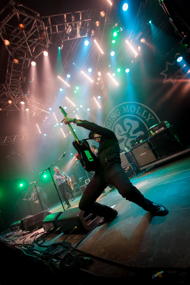 Ein musikalisches Konzert mit drei Performern auf der Bühne: zwei Gitarristen rechts mit Mikrofonen davor, ein Hintergrund mit 'MOLLEY' und zwei Schlangensymbolen sowie Scheinwerfer darüber.