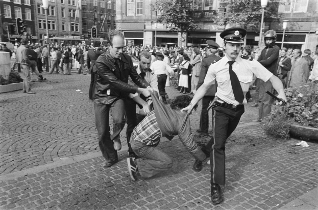 Schwarz-weißes Bild eines Mannes, der von zwei Polizisten auf einer Straße mit Zuschauern, Gebäuden, Bäumen und Verkehrsampeln im Hintergrund und einem Mann in Schuhen im Vordergrund gezogen wird.