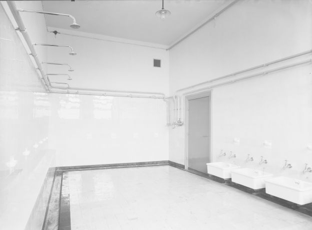 Ein Schwarz-Weiß-Foto einer öffentlichen Toilette mit Waschbecken, Hähnen, Wänden, einer Tür, sichtbaren Rohren und einer Deckenlampe.