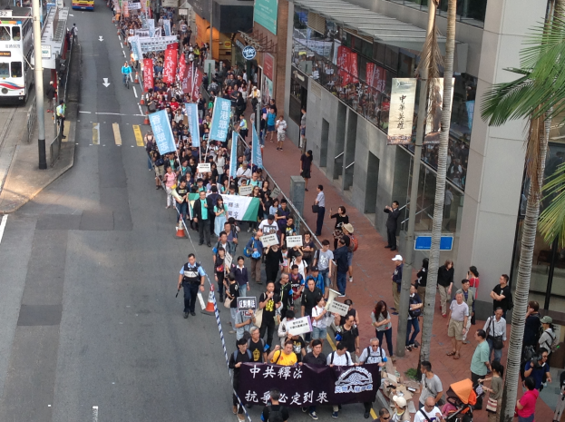 Eine große Gruppe von Menschen marschiert auf einer Straße in Hong Kong, hält Schilder und Plakate hoch, mit Bäumen, glaswandigen Gebäuden, Fahrzeugen und Schildern im Hintergrund.