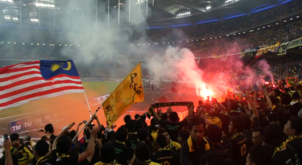 Eine große Menschenmenge in einem Stadion mit Fackeln, Fahnen und einer Kamera, mit Rauch und Schildern mit der Aufschrift "Malaysia vs Malaysia Live-Streaming" im Hintergrund.