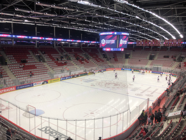 Ein Eishockeyspiel in einer großen Arena mit Zuschauern auf Sitzen und Stehplätzen, einem Zaun um die Eisfläche, einem Display mit der Aufschrift "St. Louis Blues vs Detroit Red Wings", Metallrahmen und einer Decke mit Lampen.