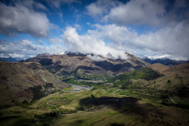 Panoramischer Blick von einem Berg in Queenstown, Neuseeland, mit grünem Gras, verstreuten Bäumen, einer gewundenen Straße und einem Himmel voller weißer, flauschiger Wolken.