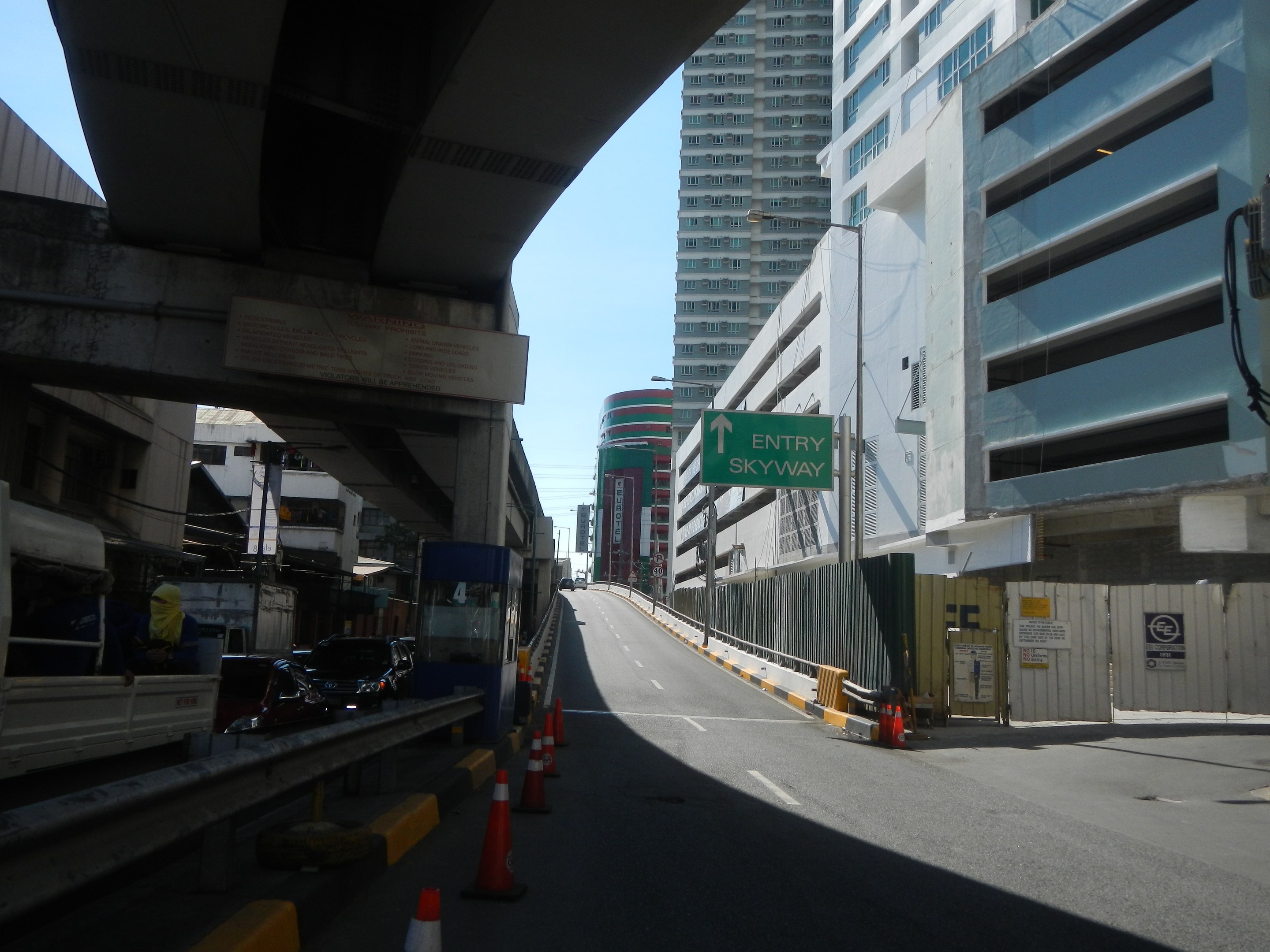 Stadtstraße mit Verkehrskegeln, Fahrzeugen, einer Brücke auf der linken Seite, Gebäuden mit Namensschildern, Laternenpfählen und einem Himmel als Hintergrund.