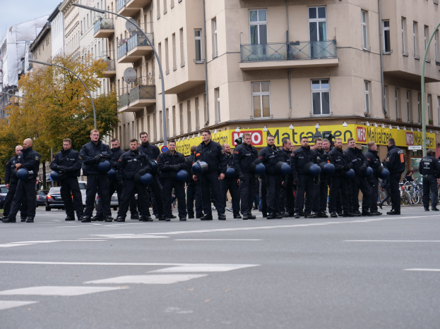 Eine Gruppe von Polizisten in schwarzen Uniformen mit blauen Helmen steht in einer Straße, die von Laternenpfählen, Bäumen und Glasfenster-Gebäuden gesäumt ist, unter einem klaren blauen Himmel.
