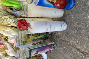 Eine improvisierte Gedenkstätte am Straßenrand mit Glasflaschen, die Fotos und Texte zeigen, einem Blumenstrauß und brennenden Kerzen auf dem Boden.