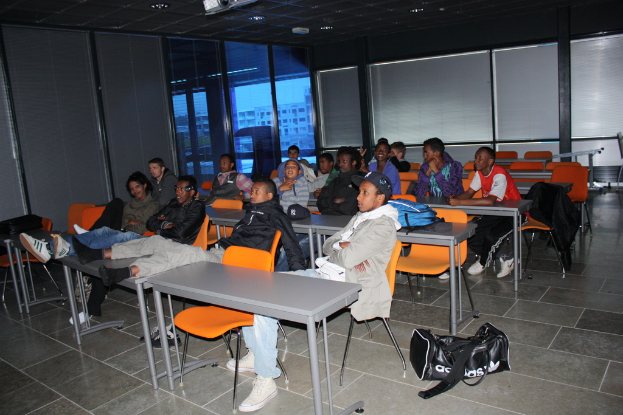 Eine Gruppe von Menschen, die auf Stühlen in einem Klassenzimmer sitzen und auf einen Schreibtisch blicken, mit Fenstern und Vorhängen an der hinteren Wand.