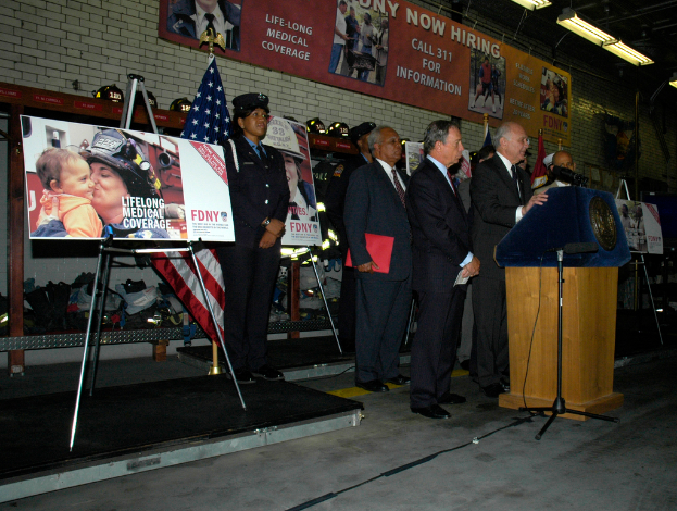 Eine Gruppe von Menschen steht in der Nähe eines Podiums mit Mikrofonen, umgeben von Tafeln auf Ständern, Flaggen und Tafeln an der Wand, alles unter hellen Lichtern.