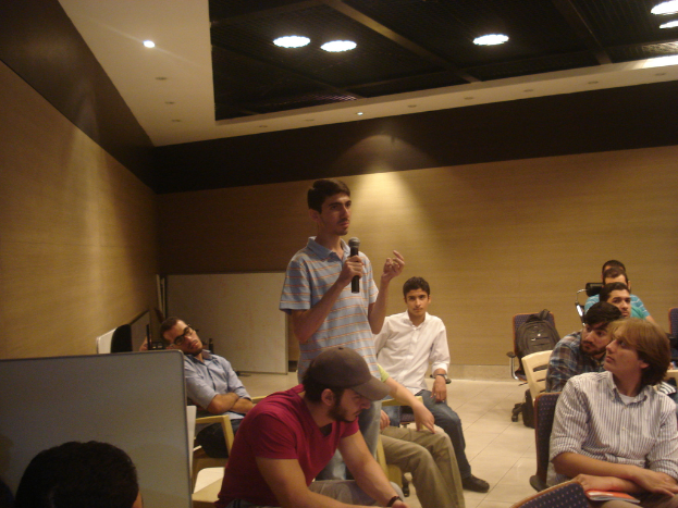 Eine Person steht mit einem Mikrofon und spricht zu einer Gruppe von sitzenden Personen in einem Büroraum.