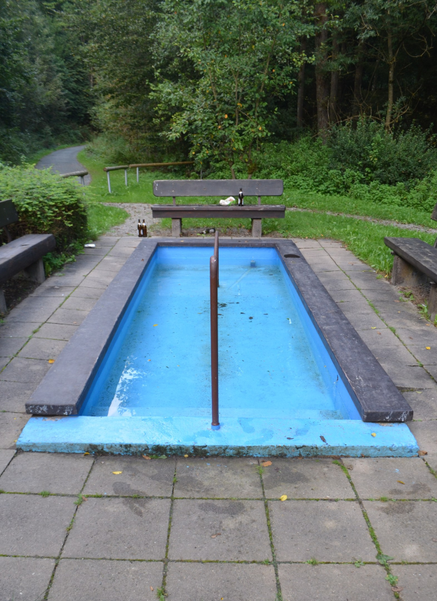Kleiner Parkpool mit Wasser, zentralem Pfahl, umgeben von Bänken, Gras, Bäumen und einer Straße.
