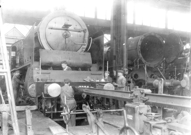 Schwarzes und weißes Foto von Männern bei der Arbeit an einem Zug in einer Fabrik, einige halten Werkzeuge, mit einer Leiter auf der linken Seite und einer Hütte im Hintergrund.