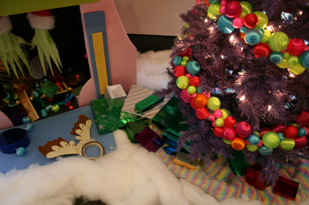 Drinnen steht rechts ein geschmückter Weihnachtsbaum mit Lichterketten und Schmuck, in der Mitte Geschenkboxen und Dekorationen, in der oberen linken Ecke eine Hütte mit Dekorationen und unten Wolle und Baumwolle.