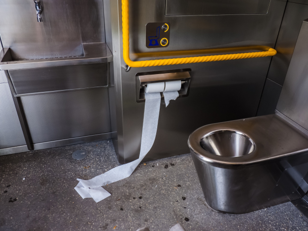 Stahltoilette mit einer Rolle Toilettenpapier daneben, einem Waschbecken mit Wasserhahn und diversen Gegenständen auf dem Boden.