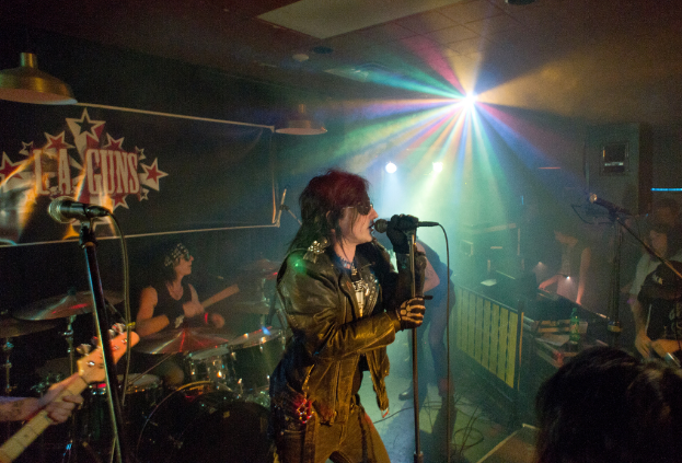 Eine Frau in einer schwarzen Jacke singt in ein Mikrofon auf der Bühne, mit einem Mann in Sonnenbrille und Stirnbinde, der ein Instrument spielt, im Hintergrund und einem Plakat mit der Aufschrift "LA Guns" im Hintergrund, unter Diskoleuchten und hängenden Lichtern.