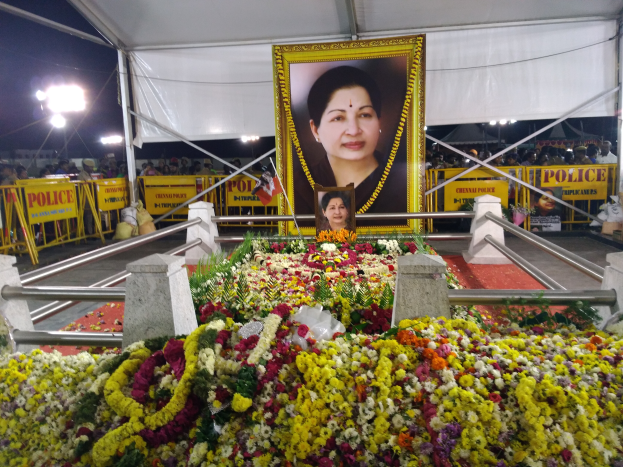 Ein Foto von einer Frau umgeben von Blumen vor einer Menge, mit Geländern, Schildern mit der Aufschrift "Sushma Swarajs Beerdigung in Hyderabad", Lichtern und einem Zelt im Hintergrund.