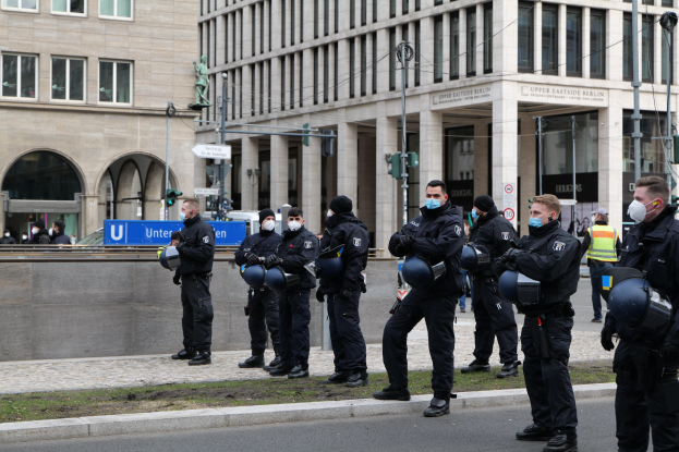 Eine Gruppe von Polizisten in schwarzen Uniformen und Masken steht vor einem Berliner Gebäude mit Glasfenstern, Säulen und Gras, einige halten Helme, mit Laternen, Verkehrsampeln, Schildern mit Text, einer Statue und anderen Menschen im Hintergrund.