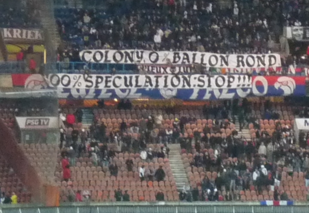 Eine große Menschenmenge in einem Stadion, mit Bannern und Stühlen im Vordergrund, einem Zaun unten und einer Treppe auf der linken Seite.