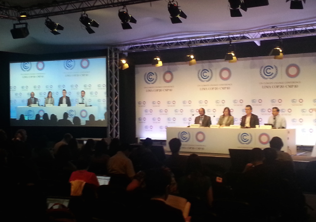 Menschen sitzen auf Stühlen auf einer Bühne mit Namensschildern und Mikrofonen auf einem Tisch, während Zuschauer von ihren Sitzplätzen aus zusehen; ein Projektionsschirm und Beleuchtung an Eisenstangen sind im Hintergrund.