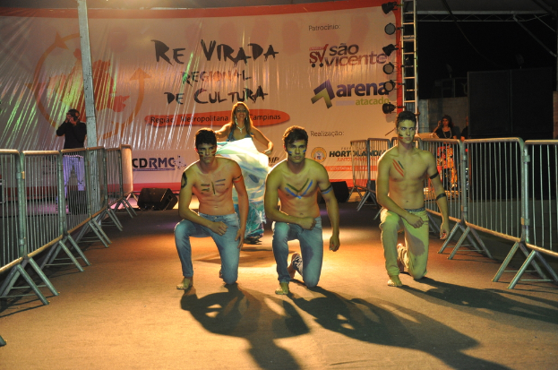 Drei shirtlose Jungs knien auf dem Boden während eines kulturellen Ereignisses, mit Grills auf beiden Seiten, einem großen Banner im Hintergrund und einer Person, die links ein Foto macht.