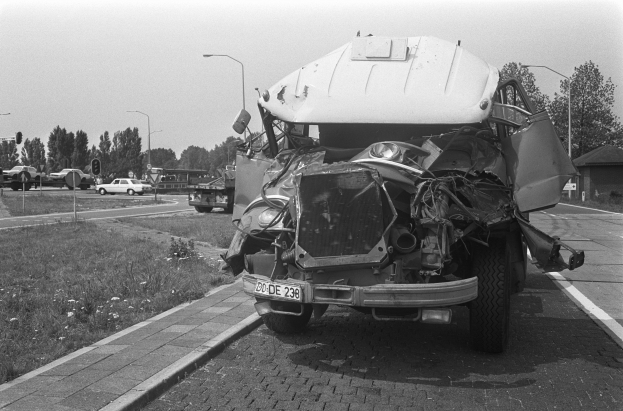 Ein Schwarz-Weiß-Foto eines schwer beschädigten Autos am Straßenrand, mit eingedellter Motorhaube, zersplitterten Fenstern und anderen Fahrzeugen, Laternenpfählen, Bäumen und einem Haus im Hintergrund.