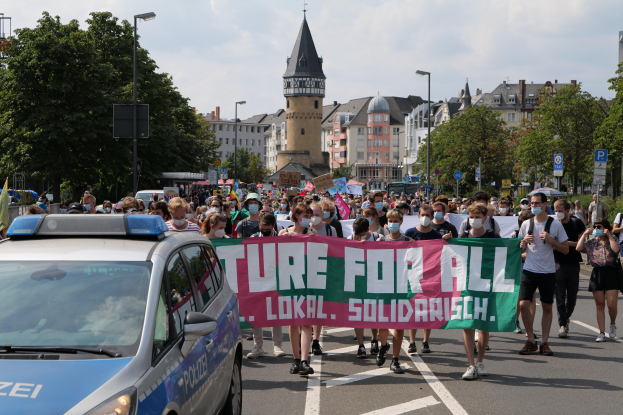 Eine große Gruppe von Menschen marschiert mit einem "Lokal Solidarität"-Schild die Straße entlang, während ein Polizeiwagen daneben geparkt ist, Bäume und Gebäude sind im Hintergrund zu sehen.