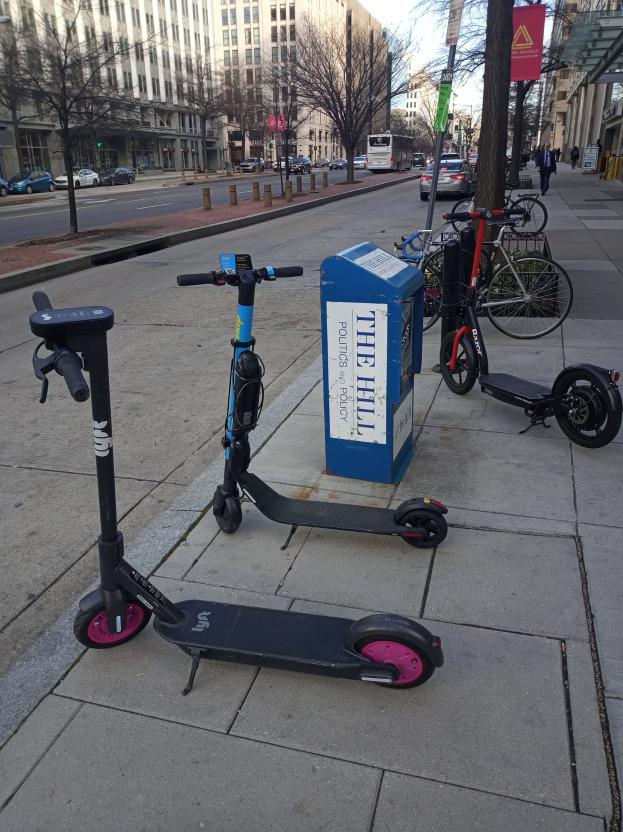 Zwei Elektro-Scooter auf einer Stadtstraße mit Fahrrädern, einem Müllcontainer und Fahrzeugen im Hintergrund.