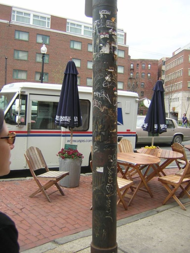Ein Tisch und Stühle sind auf einem Gehweg aufgestellt, hinter ihnen parken Autos und Lieferwagen auf der Straße, und im Hintergrund sind Gebäude zu sehen.