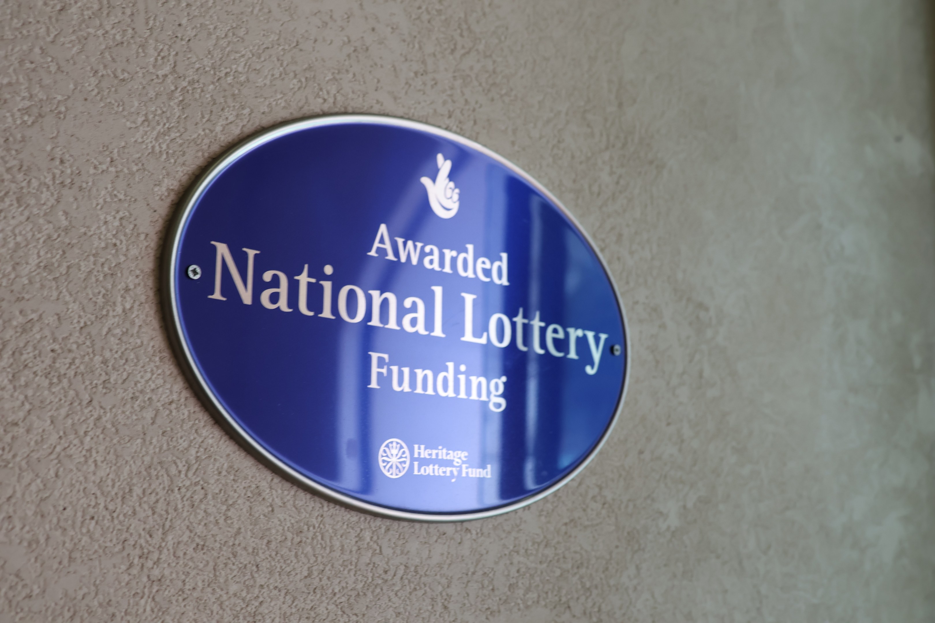 Blauer rechteckiger Schild mit weißem Hintergrund und schwarzer fetter Schrift, auf dem "National Lottery Funding" steht.
