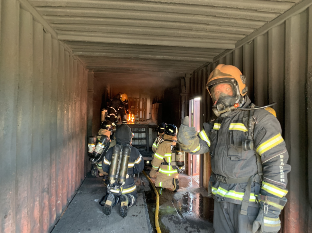 Feuerwehrleute in Schutzausrüstung innerhalb eines Containers während eines Löscheinsatzes, einer hält ein Rohr, während andere die Ausrüstung einschließlich eines Feuerlöschers verwalten.