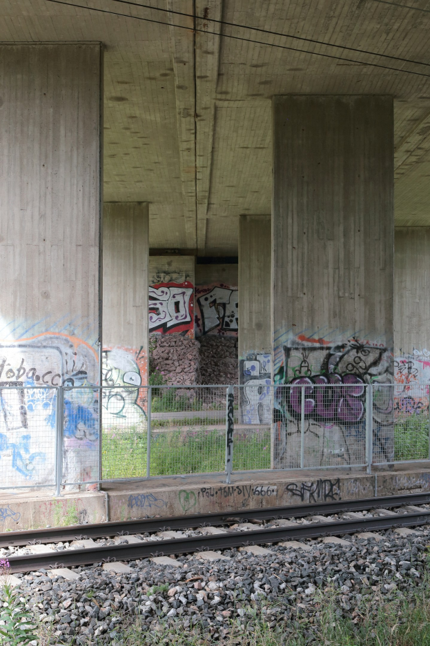 Eisenbahnschiene mit Graffiti an einer Betonwand, umgeben von Steinen, Gras, einem Zaun, Pfeilern, Überlandleitungen und einem Dach.