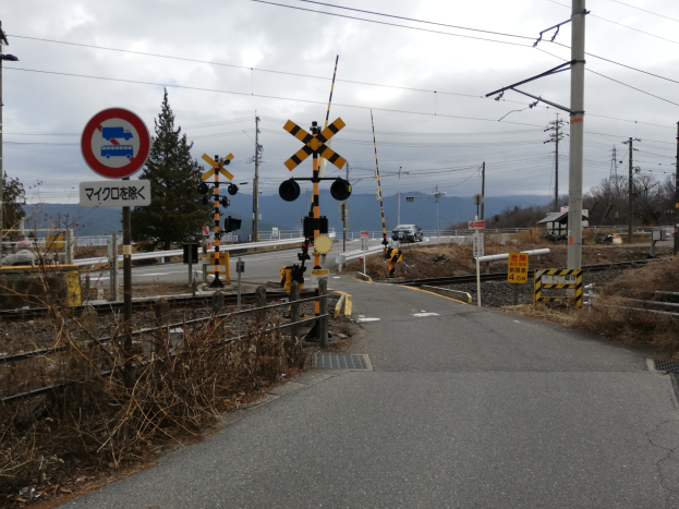 Straße mit einem Bahnübergangsschild, Eisenbahnschienen, Strommasten mit Drähten, Schilder, Fahrzeuge, Bäume, Hügel und einen bewölkten Himmel im Hintergrund.