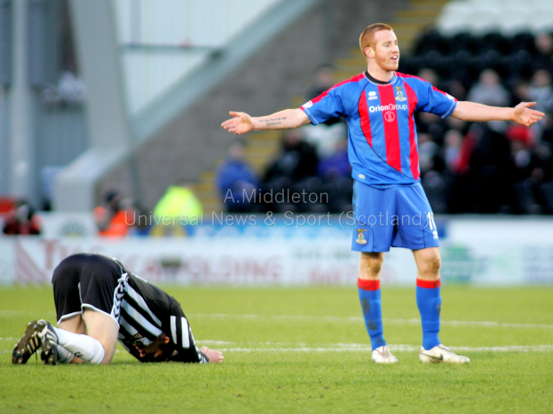 Ein Stadion mit zwei Personen im Vordergrund, eine stehend und eine kniend auf dem Gras, und einer Gruppe von Menschen im Hintergrund hinter Schildern mit Text.