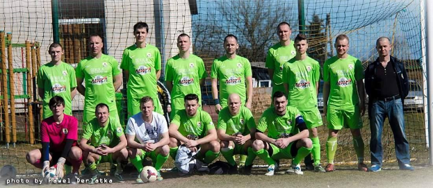 Eine Gruppe von Männern in grünen T-Shirts steht auf einem Fußballfeld neben einem Torpfosten, einige hocken, mit einem Ball vor ihnen und Gebäuden, Bäumen und Fahrzeugen im Hintergrund.