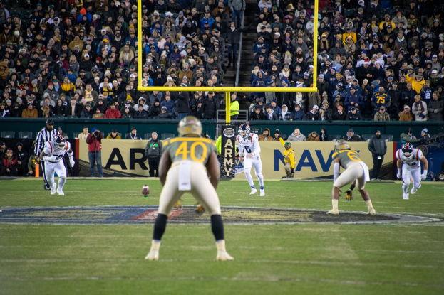 Ein Fußballspiel im Gange mit Spielern in Helmen und Sportbekleidung auf dem Feld und einer Zuschauermenge im Hintergrund, mit sichtbaren Pfählen und Tafeln mit Text.