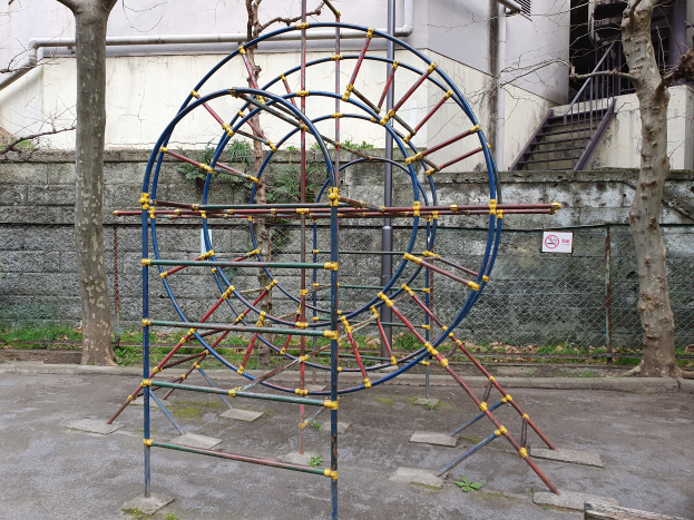 Ein Spielplatz mit einer großen Metallstruktur in der Mitte, umgeben von Bäumen und einer Wand mit Maschendrahtzaun, mit Treppen mit Geländern, die zu einem Gebäude mit Fenstern und Rohren führen, und einem Schild an einem Pfahl, alles auf einem grasbewachsenen Grund.