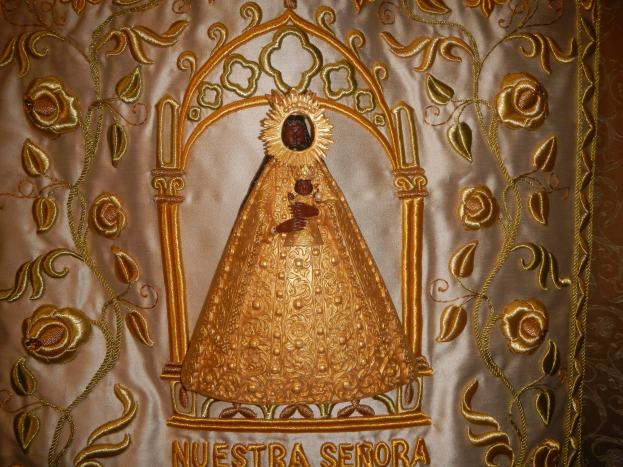 Goldbestickte Chasuble mit einer serenitätsstrahlenden Frau in einem hochgeschlossenen, langärmeligen Kleid mit einem Dutt, umgeben von detaillierten Designs und prunkvollen Texten in einem goldenen Rahmen.
