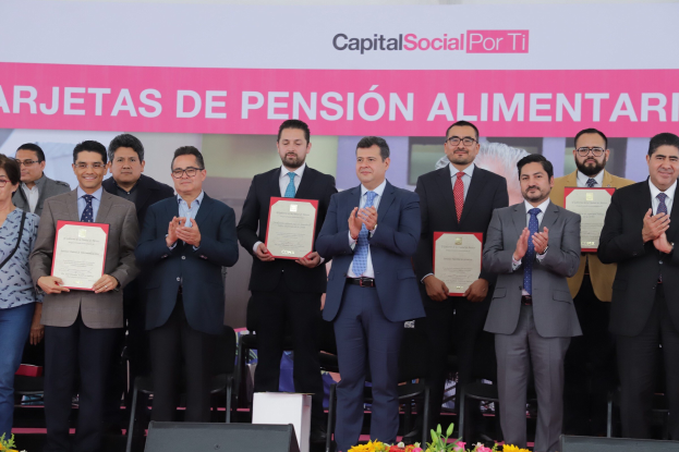 Gruppe von Menschen auf einer Bühne klappend und Zertifikate haltend, mit Stühlen in einem Halbkreis angeordnet, einem Banner mit der Aufschrift "Capital Social for Ti" im Hintergrund und Blumenbouquets und Lautsprechern unten.