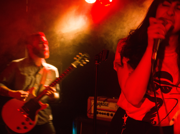 Eine Frau singt in ein Mikrofon auf der Bühne, wöhrend ein Mann im Hintergrund Gitarre spielt, mit Deckenleuchten, die die lebendige Szenerie erhellen.